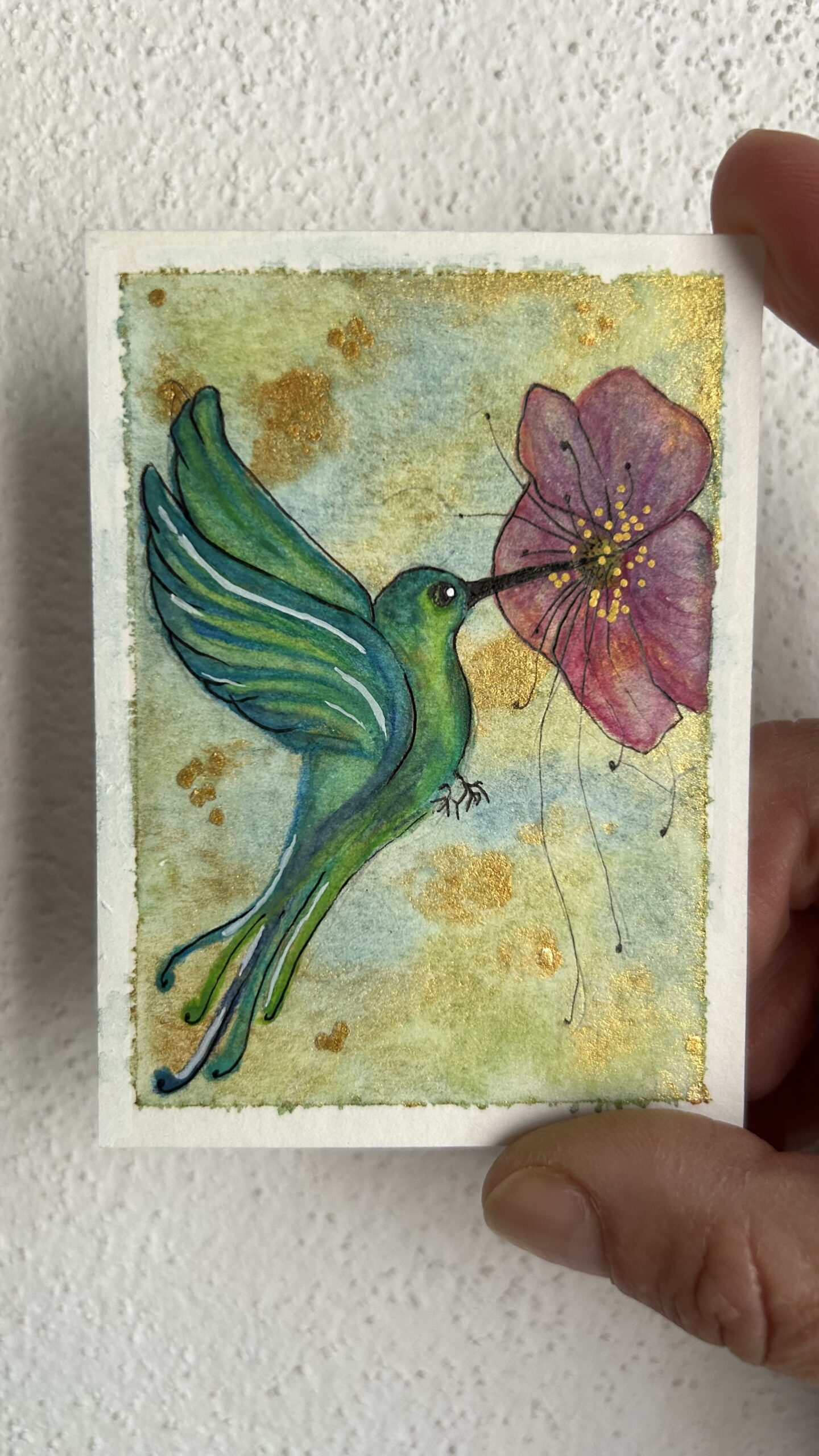 Original miniature painting “The Hummingbird Alchemist” by Nia Dannai, green hummingbird drinking nectar from hibiscus-like flower on metallic gold background, mixed media